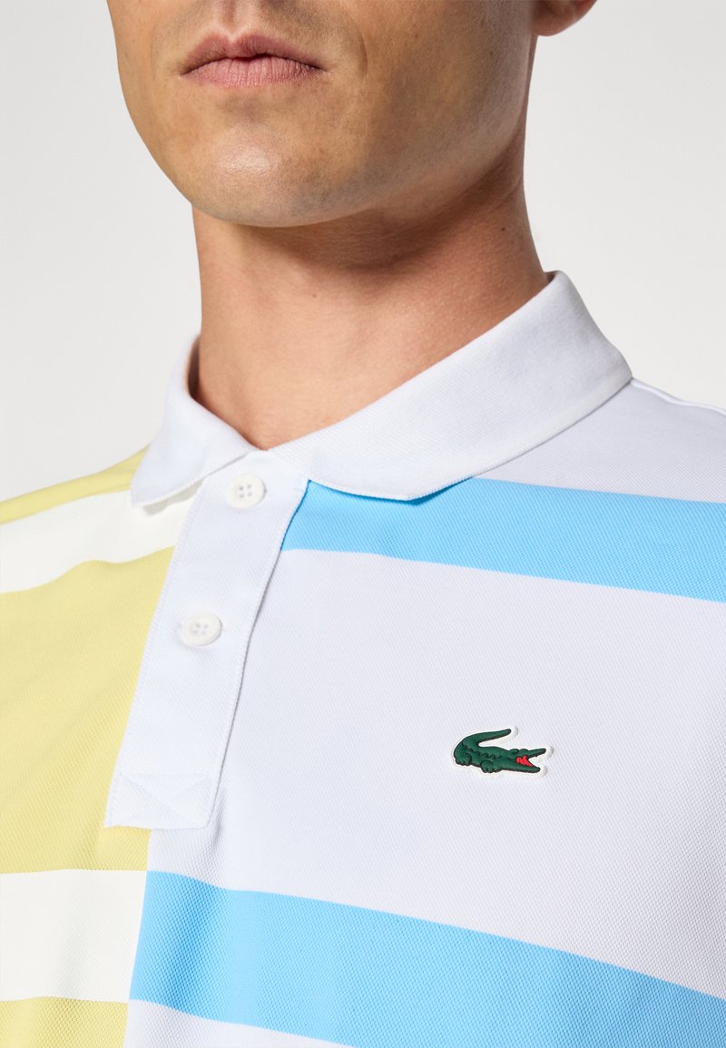 Le polo présente des rayures horizontales en jaune, blanc et bleu. Fabriqué en tissu respirant, il comprend un logo brodé vert sur la poitrine.