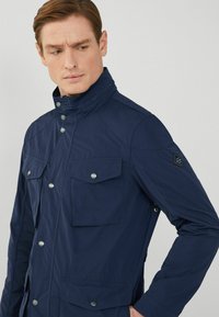 Marinblå jacka i lätt material med hög krage, två bröstfickor, tryckknappar och axeldetaljer.