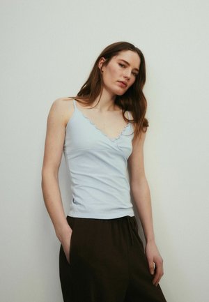 Kvinde med langt brunt hår iført en lyseblå ærmeløs top og mørke bukser, stående mod en ensfarvet lys væg.