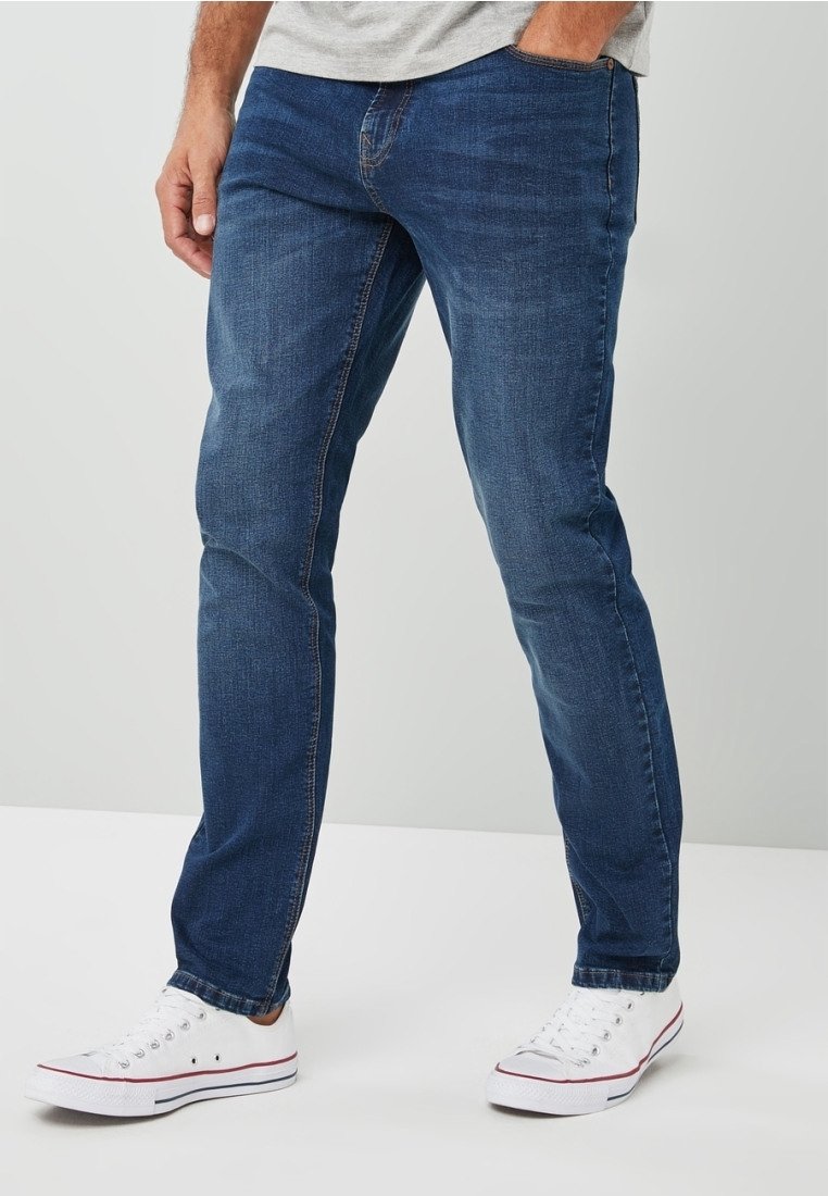 Homme portant un jean slim bleu et des baskets blanches basses, debout devant un fond uni.