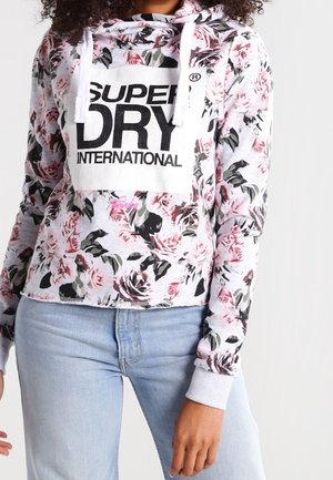 Kobieta nosząca kwiecistą bluzę z kapturem i długim rękawem z napisem „Superdry International” oraz jasnoniebieskie dżinsy, pokazana od ramion do bioder.