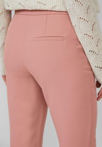 Close-up van een persoon in roze aansluitende broek en een crèmekleurige gebreide trui met textuurpatronen op de mouwen.