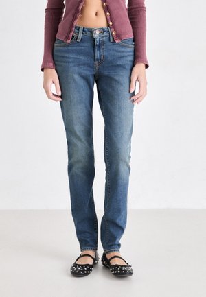 Frau trägt hoch taillierte blaue Jeans, ein bauchfreies, mauvefarbenes, geripptes Hemd mit Knöpfen und schwarze flache Schuhe mit Nieten vor einem schlichten hellen Hintergrund.