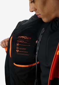 Giacca nera con accenti arancioni, realizzata in materiale impermeabile Exo Shield, con una classificazione impermeabile di 30k, elasticità a 4 vie e dettagli isolanti per il calore.