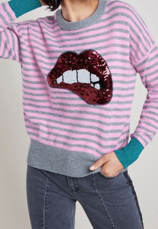 Femme portant un pull rayé rose et gris avec des poignets bleu sarcelle, arborant un grand motif de lèvres rouges à sequins sur le devant.