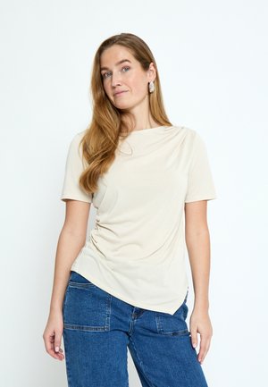 Vrouw met lang lichtbruin haar, gekleed in een crèmekleurige top met korte mouwen en een blauwe spijkerbroek, staand tegen een effen witte achtergrond.