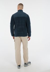 Man die met zijn rug naar de camera staat, gekleed in een marineblauwe fleecejack, beige cargobroek en grijze en zwarte wandelschoenen op een witte achtergrond.