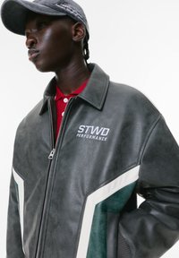 Giacca in ecopelle grigia con accenti bianchi e verdi, dotata di cerniera frontale, colletto e logo "STWD PERFORMANCE" sul petto.