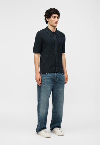 Άνδρας στέκεται φορώντας μαύρο πουκάμισο με κοντά μανίκια και κουμπιά, άνετο μπλε τζιν και λευκά αθλητικά παπούτσια, μπροστά σε απλό ανοιχτό γκρι φόντο.