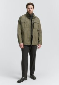 Olivgrüne Jacke mit hohem Kragen, Reißverschluss und zwei Brusttaschen. Kombiniert mit dunkelgrauen Hosen und schwarzen Schuhen.