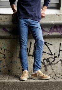 Personne portant un jean bleu, des baskets beiges et un sweat-shirt bleu marine foncé, appuyée contre un mur en béton couvert de graffiti à l'extérieur.