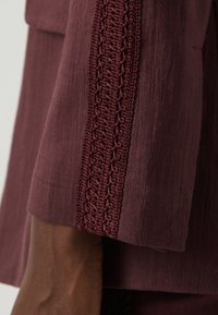 Manga de tela burdeos con detalle de crochet texturizado a lo largo de la costura, con un acabado suave y un ligero brillo.