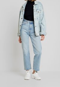 Lichtblauwe denimjacket over een zwart top, gecombineerd met high-waisted lichtblauwe jeans en witte sneakers. Casual uitstraling, lichte slijtage op de jeans.