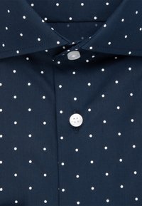 Marineblauw stoffen shirt met een button-down kraag, witte polkadots en een witte knoop bij het beleg. Gladde textuur.