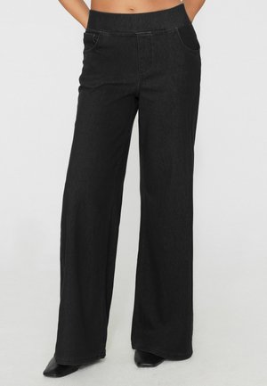Pantaloni neri a gamba larga con cintura elastica indossati con scarpe nere con punta, mostrati dalla vita ai piedi su uno sfondo semplice.