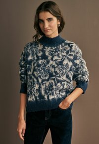 Strickpullover in Blau und Creme, mit einem hohen gerippten Kragen, lässiger Passform und strukturiertem Muster. Kombiniert mit dunklen Jeans.
