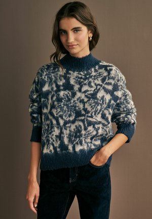 Strickpullover in Blau und Creme, mit einem hohen gerippten Kragen, lässiger Passform und strukturiertem Muster. Kombiniert mit dunklen Jeans.