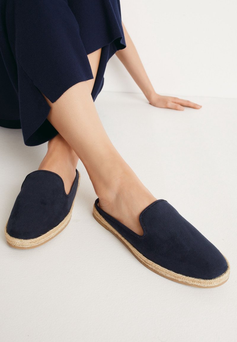 Chaussures en daim bleu marine à enfiler avec une semelle tressée en jute et une ouverture supérieure incurvée, présentant une texture lisse et un design minimaliste.