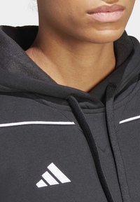 Sudadera negra con acentos blancos, con cremallera frontal y cordón, hecha de tela texturizada. Logo de Adidas exhibido en el lado izquierdo.