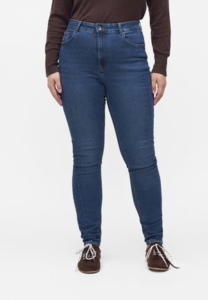 Vrouw die blauwe skinny jeans, een bruin shirt met lange mouwen en bruine schoenen met witte veters draagt, staand tegen een witte achtergrond.