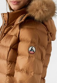 Giacca imbottita marrone con cappuccio rifinito in pelliccia, tessuto trapuntato e logo ricamato circolare sulla manica.