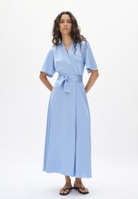 Hellblaues Wickelkleid mit kurzen Ärmeln, Bindegürtel in der Taille, schwingender Rock, aus glattem Stoff, ergänzt durch flache schwarze Sandalen.