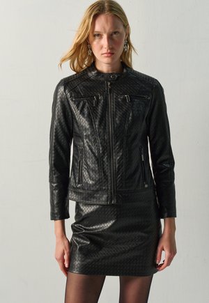Mujer rubia con una chaqueta de cuero negra texturizada con bolsillos con cremallera y una falda de cuero negra a juego sobre un fondo liso.