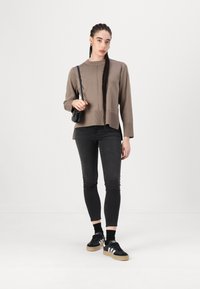 Brązowy sweter dzianinowy z długimi rękawami, czarne obcisłe dżinsy i czarne sneakersy z białymi paskami. Modelka trzyma małą czarną torebkę.