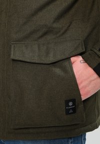 Kurtka w kolorze oliwkowej zieleni z teksturowanej tkaniny, z dużą klapową kieszenią i metką marki na dolnej lewej części. Zbliżenie na rękę w kieszeni.