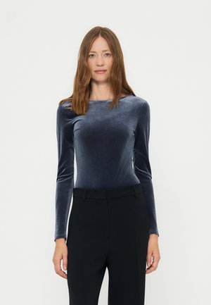 Femme aux cheveux lisses châtain clair porte un haut bleu en velours à manches longues ajusté et un pantalon noir, debout devant un fond blanc uni.
