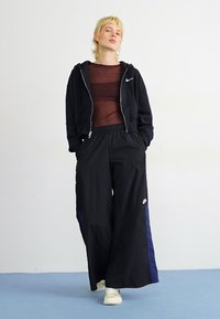 Svart zip-up hoodie med vit logotyp, transparent brun topp och lösa svarta byxor med marinblå detaljer. Vita sneakers fulländar outfiten.