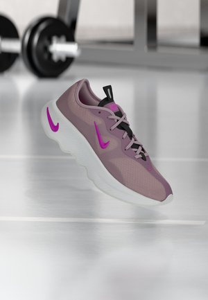 Chaussure de sport lavande et rose avec semelle blanche flottant en intérieur, poids de gym flous en arrière-plan sur un sol réfléchissant.