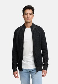 Sort zip-up cardigan med struktureret strik, raglanærmer og høj krave, båret over en hvid t-shirt og lyseblå jeans.