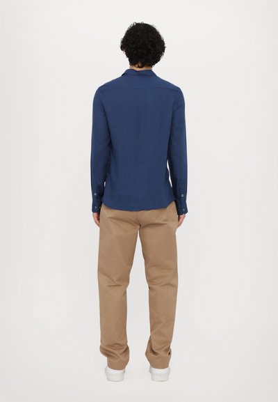 Marineblauw overhemd met lange mouwen, een kraag, knoopmanchetten en een rugpand, gecombineerd met beige broek en witte sneakers, gezien vanaf de achterkant.