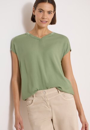 Femme portant un haut vert sauge à manches courtes et un pantalon beige, debout contre un mur clair uni.