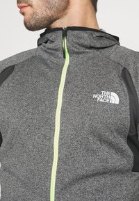 Giacca grigia con zip nera, dotata di zip verde fluo e logo "The North Face" sul petto. Design in tessuto testurizzato.