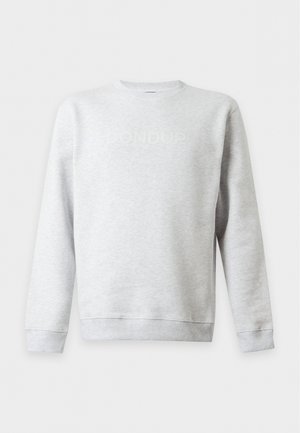 Jeune homme portant un sweat-shirt gris clair à manches longues, un jean bleu ample et des baskets noires, debout devant un fond blanc uni.