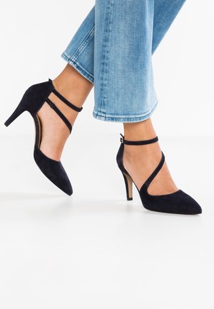 Granatowe zamszowe buty na wysokim obcasie z ostrymi czubkami i paskami na kostce. Charakteryzują się eleganckim wzornictwem i cienkimi obcasami, na białym tle.