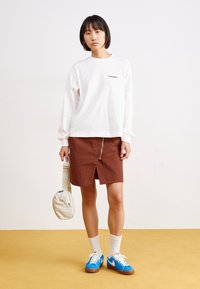 Bílá košile s dlouhým rukávem a logem, hnědá sukně na zip, modré a bílé tenisky a krémová crossbody taška na žluté podlaze.