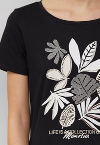 Zwart t-shirt met korte mouwen, versierd met een bladerenmotief in wit, beige en gedetailleerde zwarte omlijning; tekst luidt "HET LEVEN IS EEN VERZAMELING VAN Herinneringen."