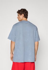 Mand, der vender væk, iført en løs, falmet blå t-shirt og klare røde shorts mod en enkel hvid baggrund.