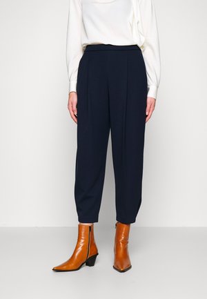Femme portant un pantalon bleu marine taille haute coupé au-dessus de la cheville avec des bottines en cuir marron à bout pointu et un chemisier blanc rentré.