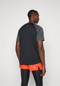 Sort atletisk t-shirt med grå raglanærmer, glat tekstur, har reflekterende detaljer på ryggen og en afslappet pasform.
