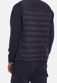 Navy puffy vest with horizontal quilted design, made of shiny material. Worn over a dark long-sleeve shirt, featuring contrasting cuffs.