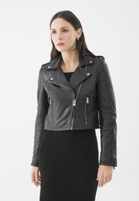 Chaqueta de moto recortada de cuero negro con cremalleras plateadas, botones a presión y hombreras, combinada con un vestido negro acanalado.