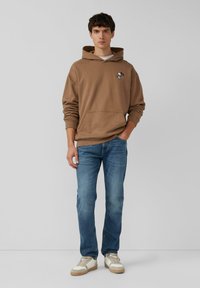 Brązowa hoodie z kieszenią i haftowanym wzorem, w połączeniu z jasnoniebieskimi dżinsami i białymi trampkami z beżowymi akcentami. Gładkie tło.