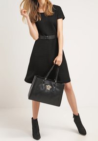 Svart texturerad klänning med korta ärmar, åtdragen i midjan med ett svart bälte. Svart tote-väska med guldlejonemblem och två handtag. Svarta ankelstövlar.