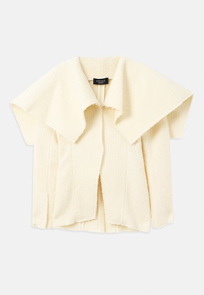 Veste crème texturée avec un large col et une coupe ouverte. Dispose de manches courtes et d'une silhouette moderne et carrée.