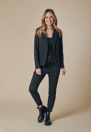 Schwarzer Blazer mit maßgeschneiderter Passform, kombiniert mit einer passenden Weste und Jogginghosen. Glatter Stoff, knöchellange Hosen und schwarze Stiefeletten.