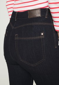 Los jeans de mezclilla oscura cuentan con un diseño ajustado, una etiqueta trasera de cuero marrón, costuras en contraste y bolsillos traseros estándar con remaches de cobre.
